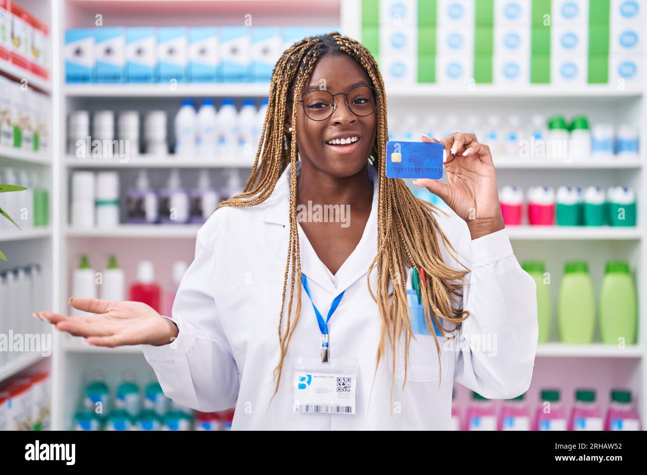 African american woman with braided hair working at pharmacy drugstore holding credit card celebrating achievement with happy smile and winner express Stock Photo