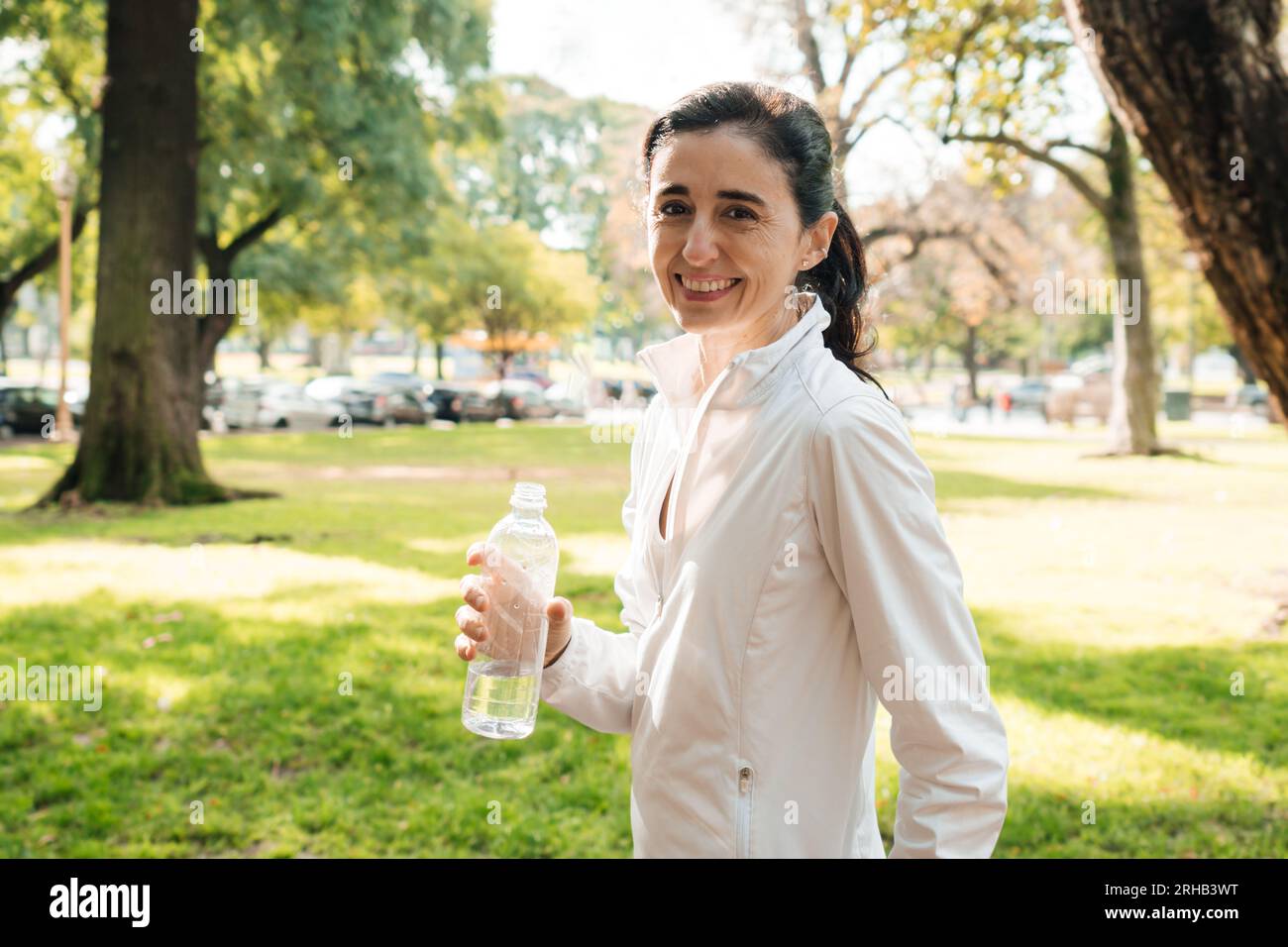 Middle aged woman smiles and looks at camera with water bottle in hand, after workout in park. Stock Photo