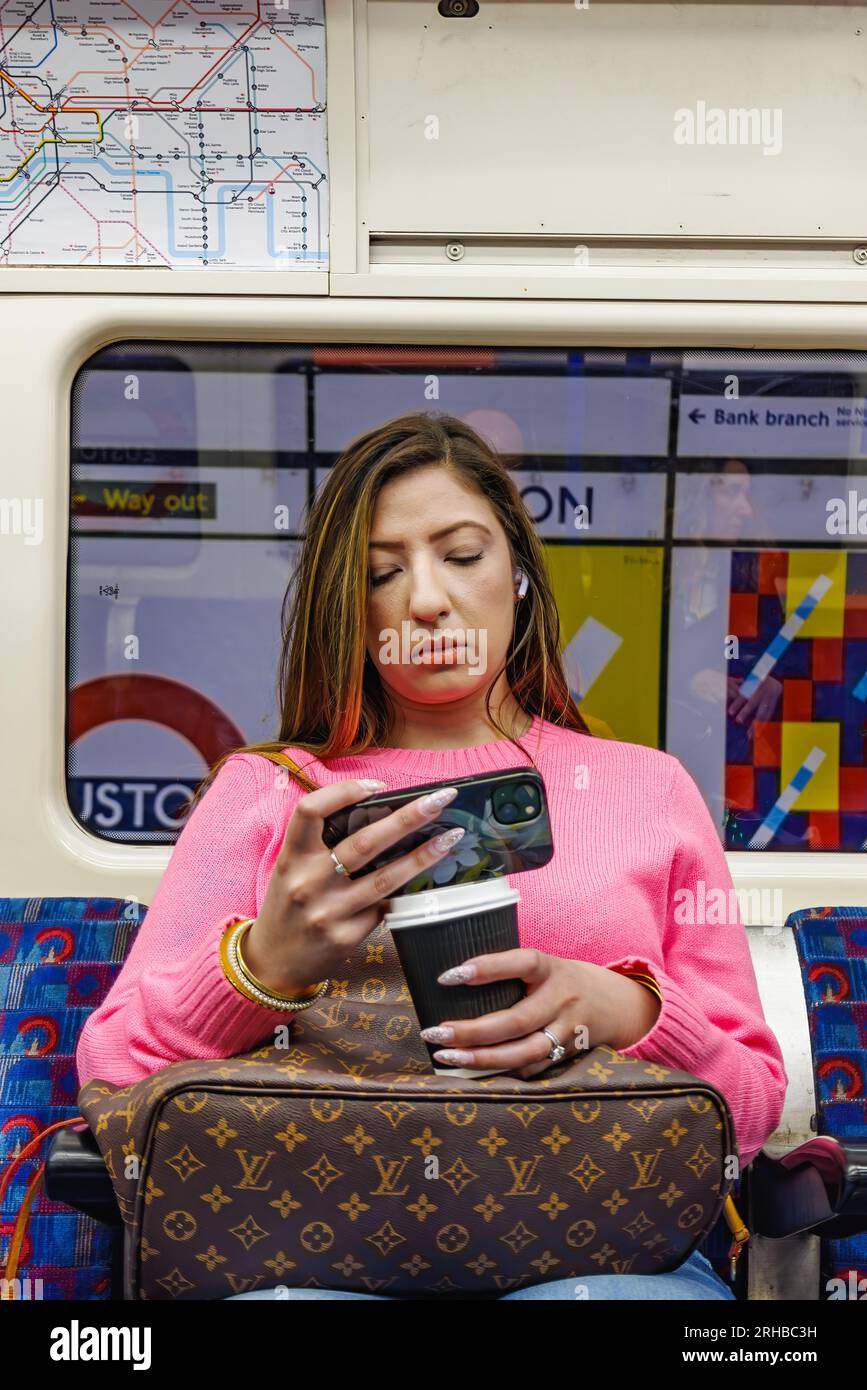 London, UK - May 23, 2023: A young woman traveling by subway is using her mobile phone while having a coffee to go and wearing a Louis Vuitton handbag Stock Photo