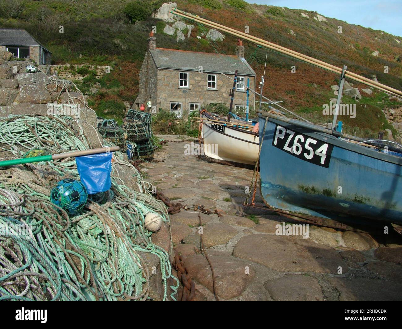 Crab and lobster pots and fishing boats above the slipway with an old cottage in the background at Penberth Cove Harbour, Cornwall, England, UK. Stock Photo