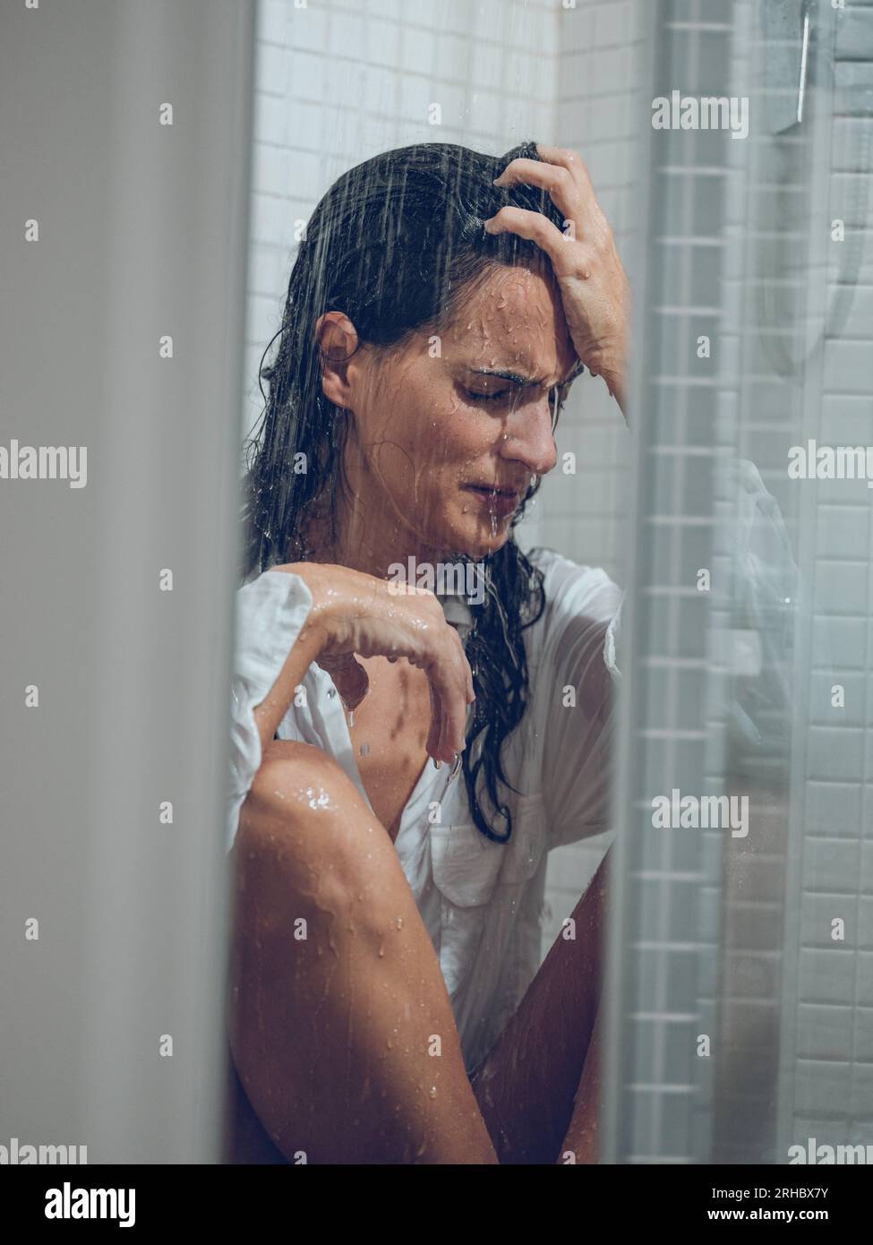 Sorrowing woman with long hair getting shower in white shirt while sitting behind transparent curtain Stock Photo