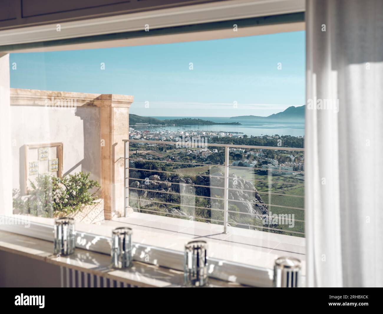 Through window view of room of hotel overlooking balcony and green terrain and azure sea in sunny day in Pollensa city of Mallorca island Stock Photo