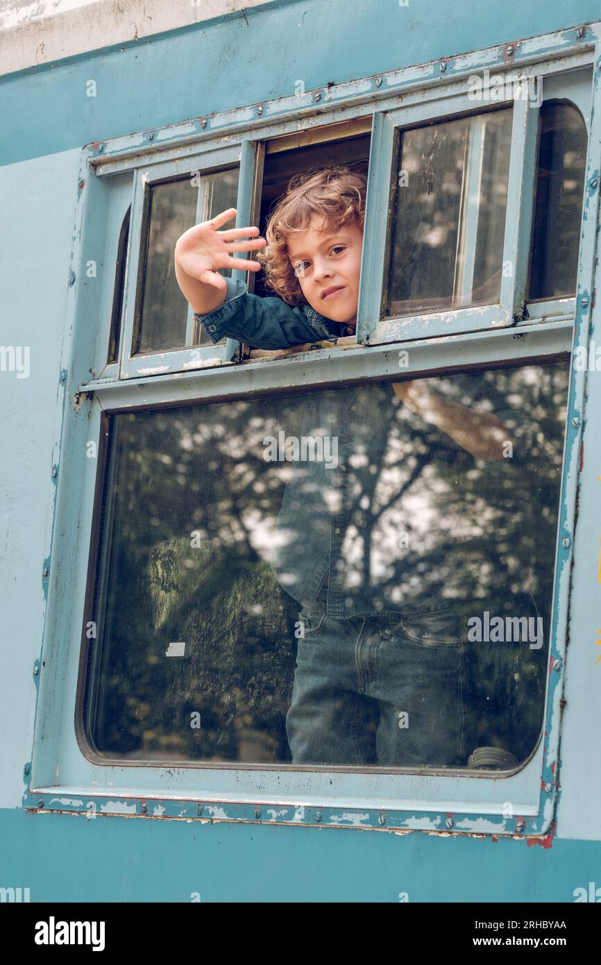 Blond haired kid looking out of opened window and waving hand while preparing for train ride and looking at camera Stock Photo