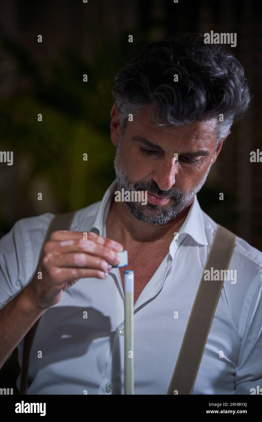 Focused bearded man in white shirt and suspenders looking down and applying chalk on cue while playing billiards in dark room on blurred background Stock Photo