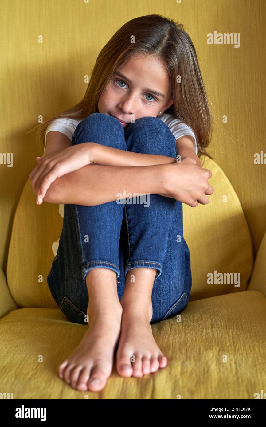 Full body of adorable barefoot child embracing knees and looking at camera while sitting in soft armchair in room Stock Photo