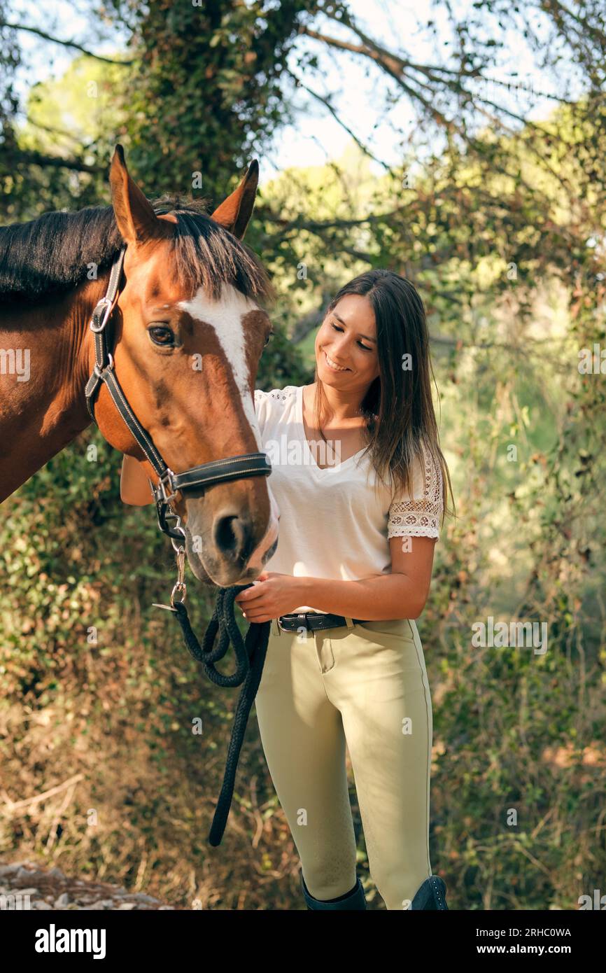 Positive female equestrian holding reins and caressing chestnut horse while standing together in woods on sunny day Stock Photo