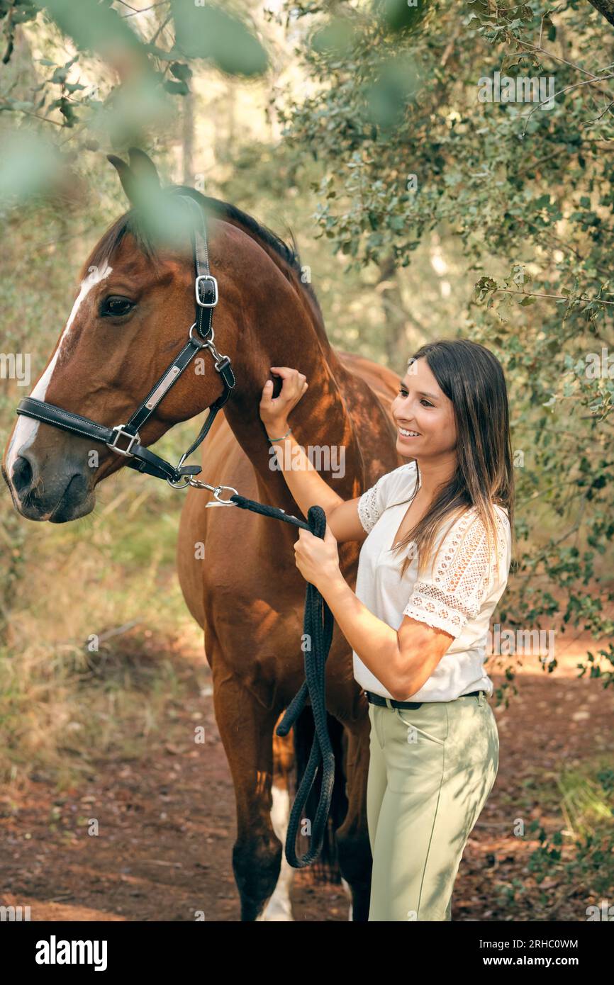 Cheerful female equestrian stroking brown horse while holding reins and standing in lush forest Stock Photo