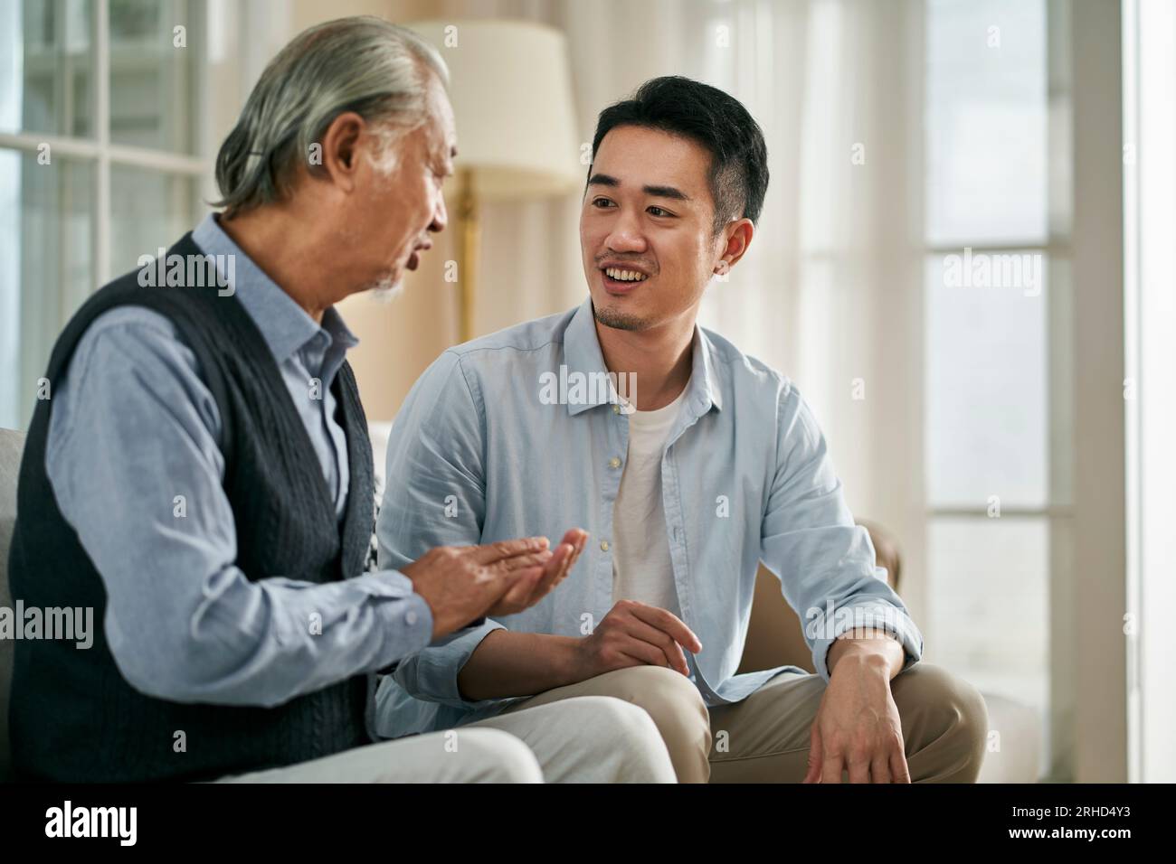 senior asian father having a good time sitting on couch at home chatting with adult son Stock Photo