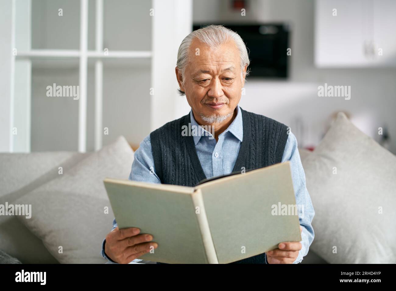senior asian man sitting on couch in living room at home reading a book Stock Photo