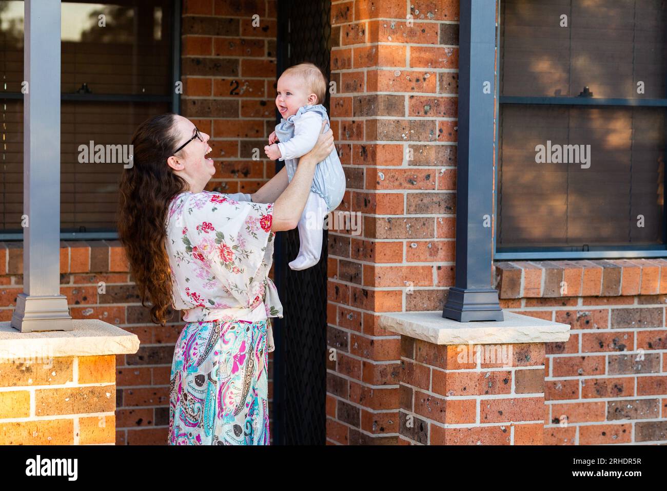 Young mum lifting up baby girl by front door of suburban home smiling together Stock Photo