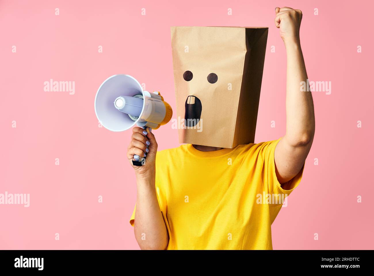 Emotional woman with paper bag on head making announcement with megaphone in hands. anonymous female screaming in loudspeaker on pink background Stock Photo