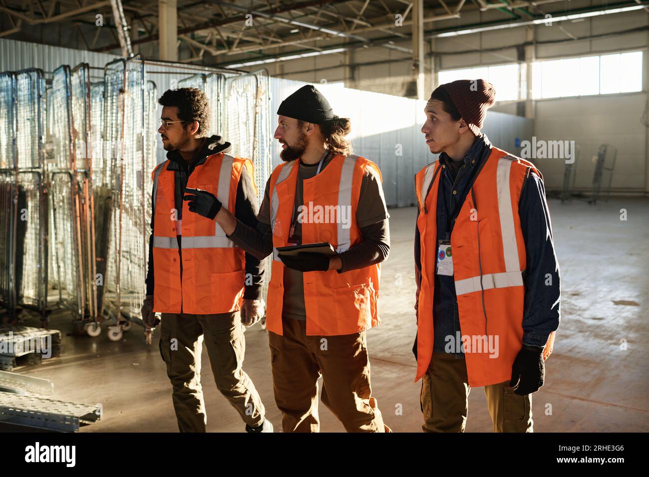 Three young intercultural builders in safety jackets walking along spacious workshop and discussing which new equipment should be acquired Stock Photo