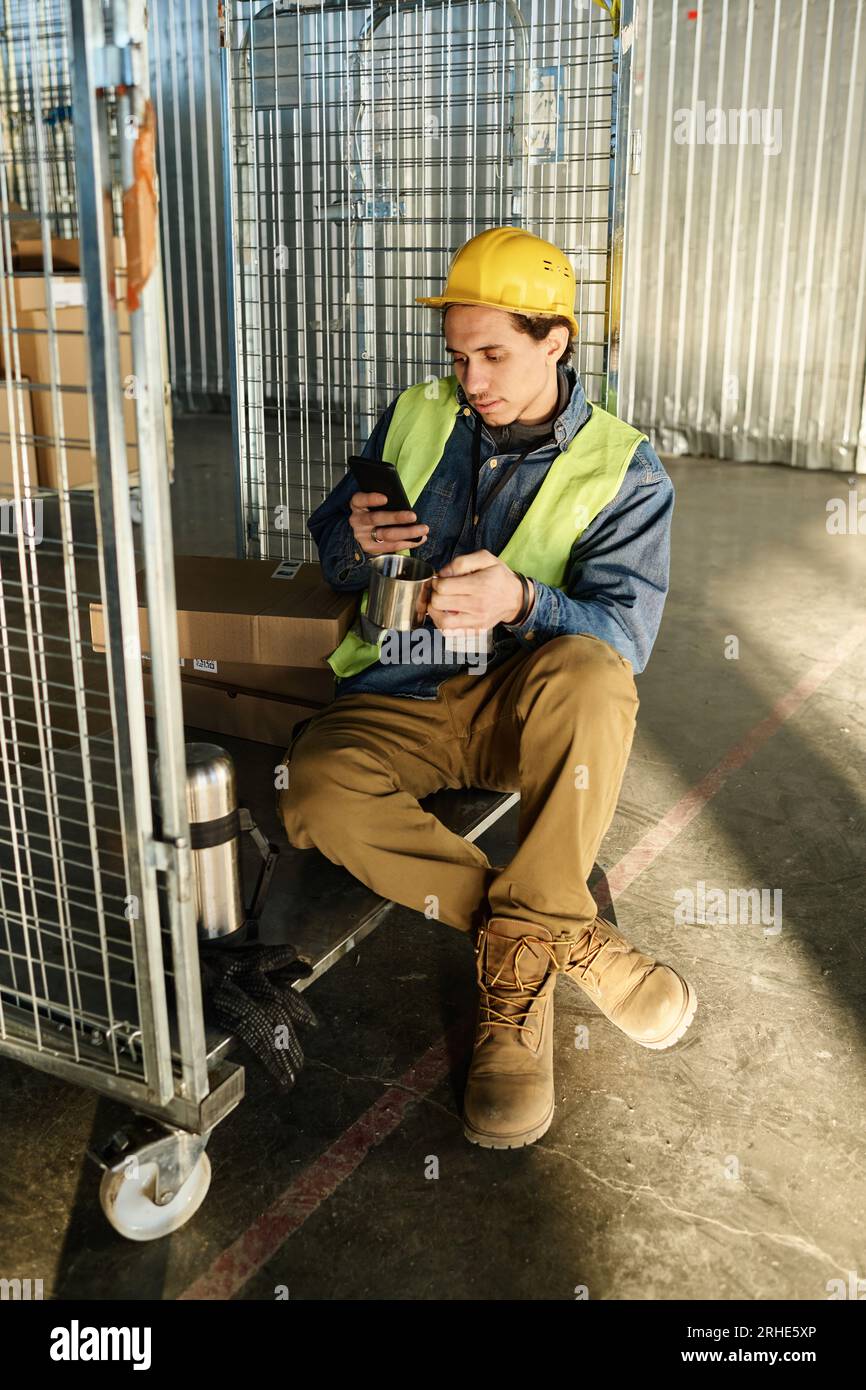 Young multiethnic male engineer in protetive workwear scrolling in mobile phone while sitting on large metallic cart and having tea at break Stock Photo