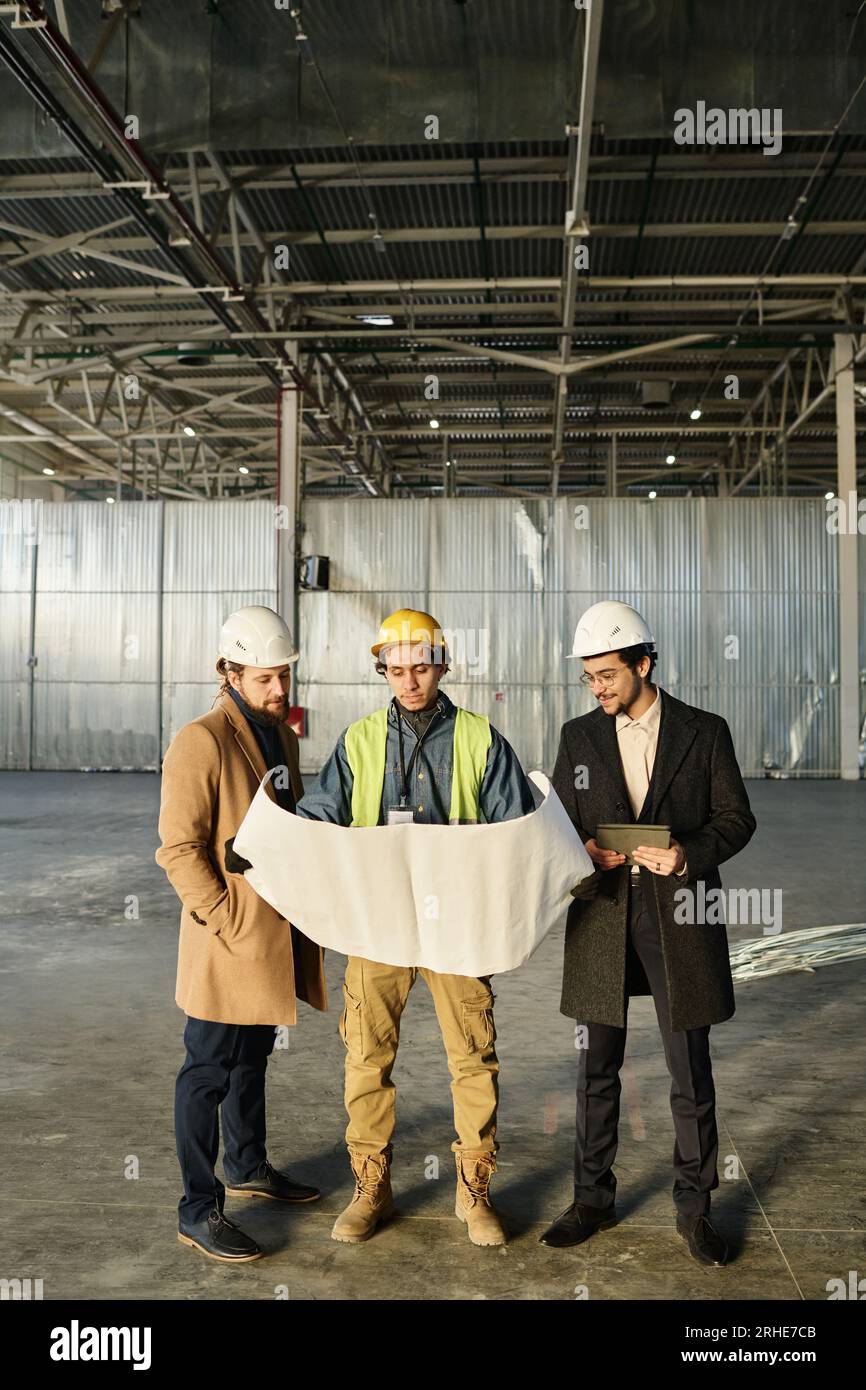 Front view of team of young architects or engineers discussing building plan or sketch on unrolled blueprint held by worker in safety vest Stock Photo