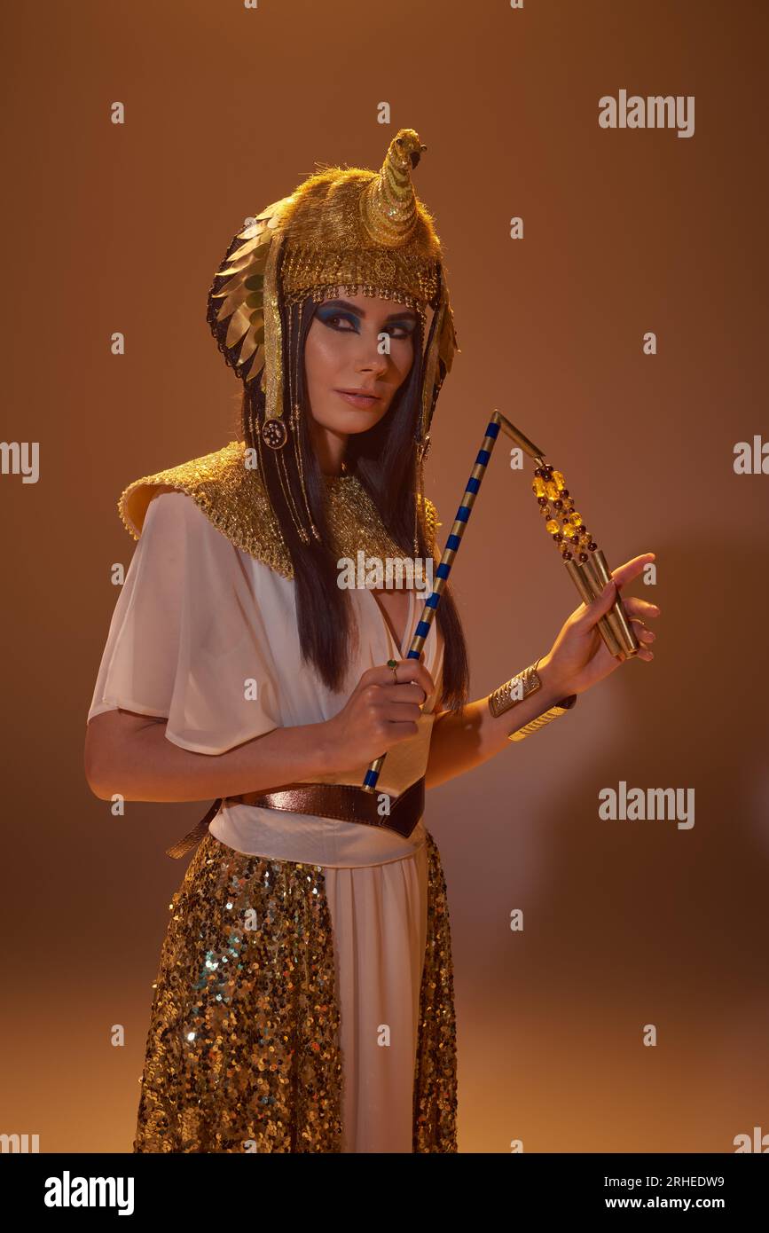 Beautiful woman in egyptian attire and headdress holding flail while posing on brown background Stock Photo