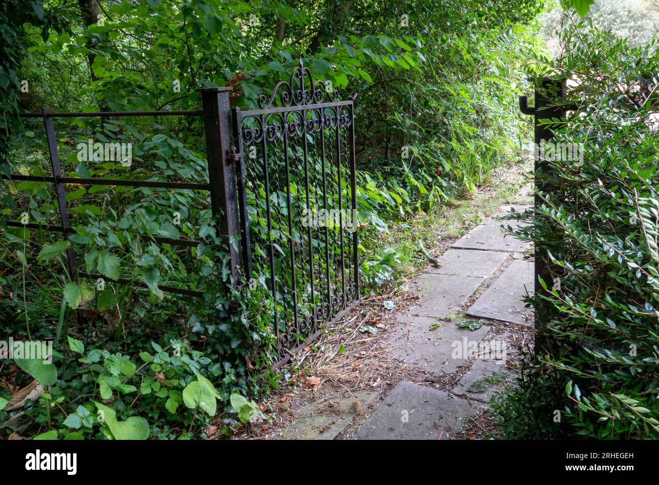 An open wrought iron garden gate allowing access to a slab pathway surrounded by rich shrubbery and trees Stock Photo