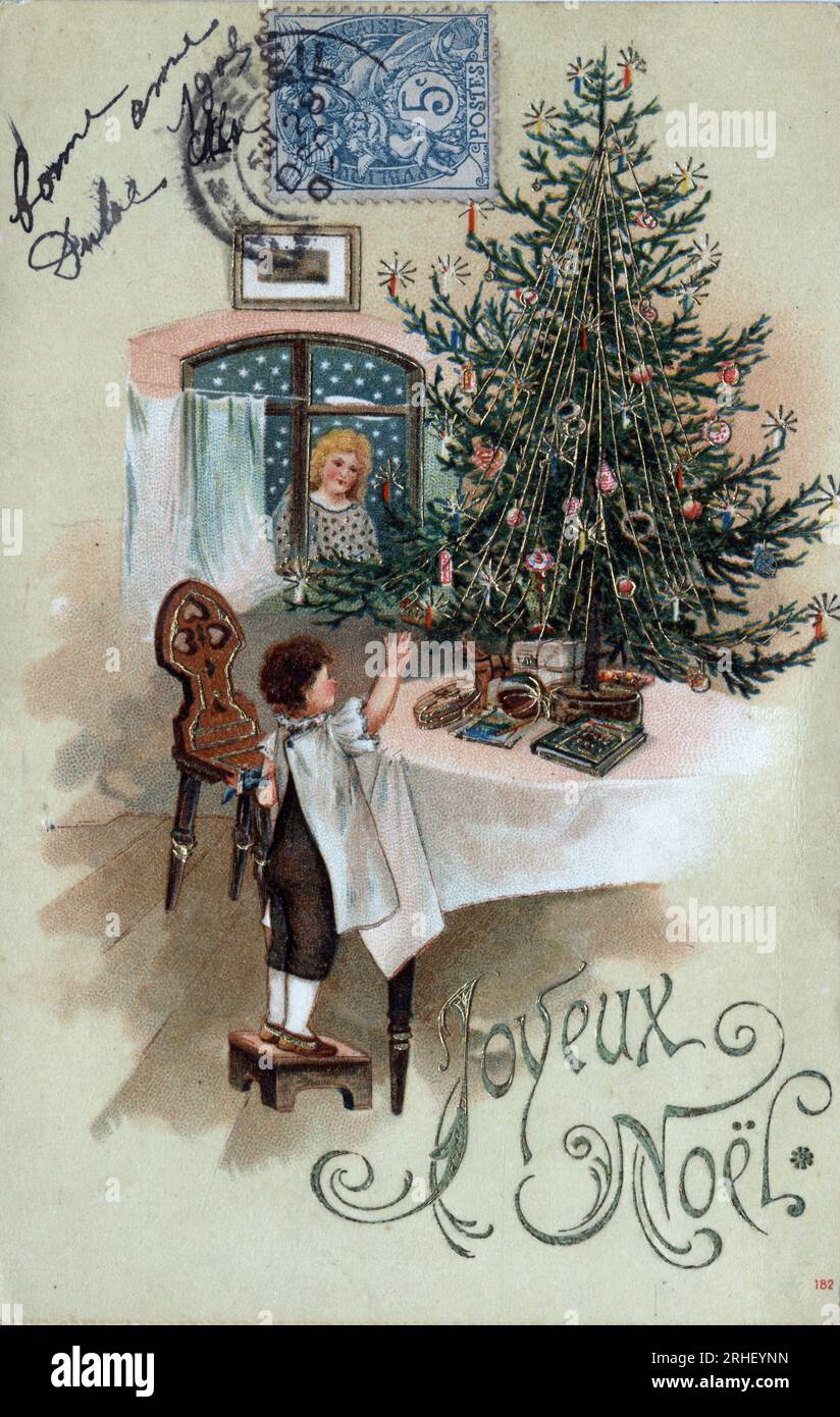 Carte de voeux pour Noel : un enfant devant un sapin de Noel pose sur une table, observe par un ange a la fenetre - Carte postale datee 1905 Stock Photo