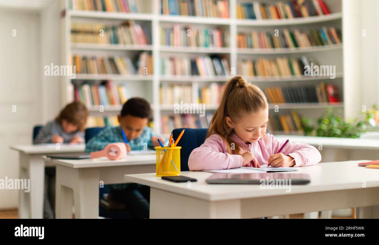 Group of diverse schoolboys and schoolgirl sitting at desks in classroom at primary school, writing in notebook Stock Photo