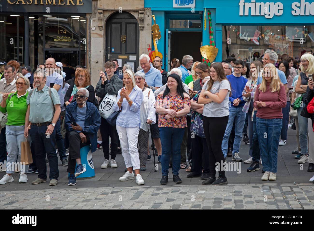 City centre, Edinburgh, Scotland, UK. 16 August 2023.  Cloudy conditions in the city centre for those visiting the Edinburgh Festival Fringe, small audiences for some street performers on the High Street. Pictured: A small audiience is entertained outside the Fring shop and office on the High Street.. Credit: Archwhite/alamy live news Stock Photo