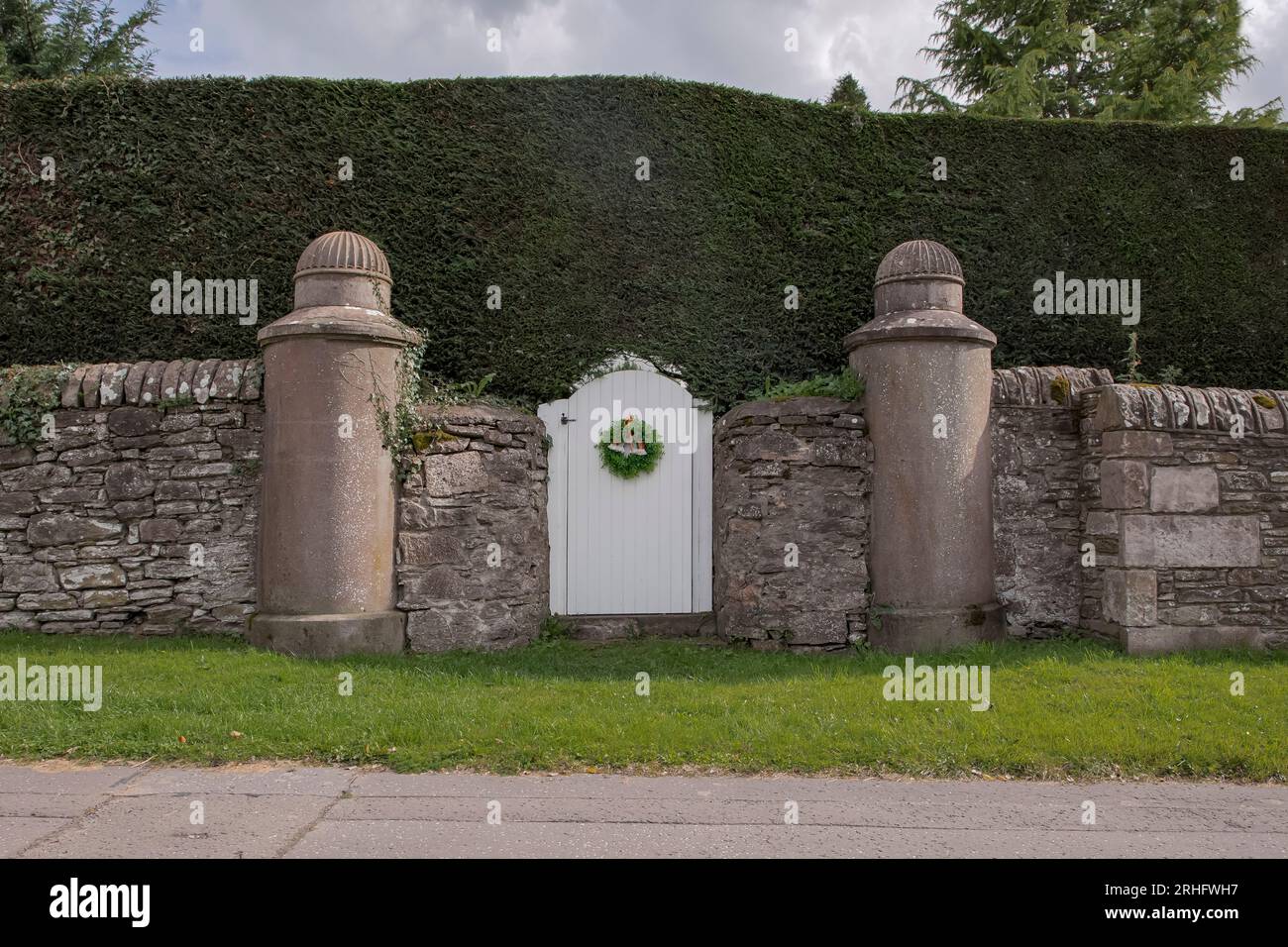 Unique white arched gate in a large conifer hedge which has a garland attached Stock Photo