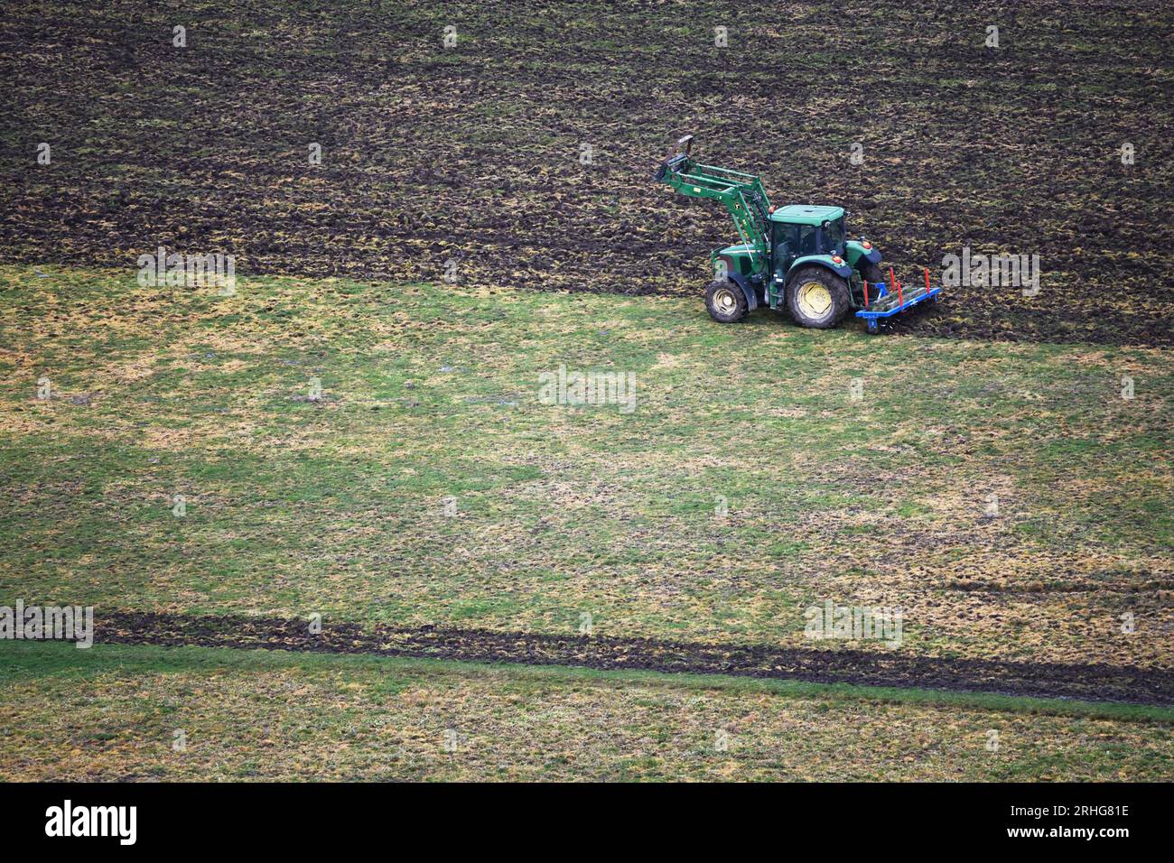 Green tractor plowing a field followed by a blue harrow, working the land efficiently. Stock Photo
