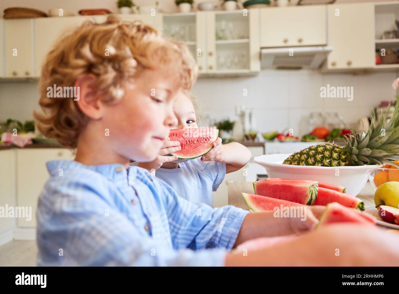 Cute girl holding fresh watermelon slice while standing with brother in kitchen at home Stock Photo