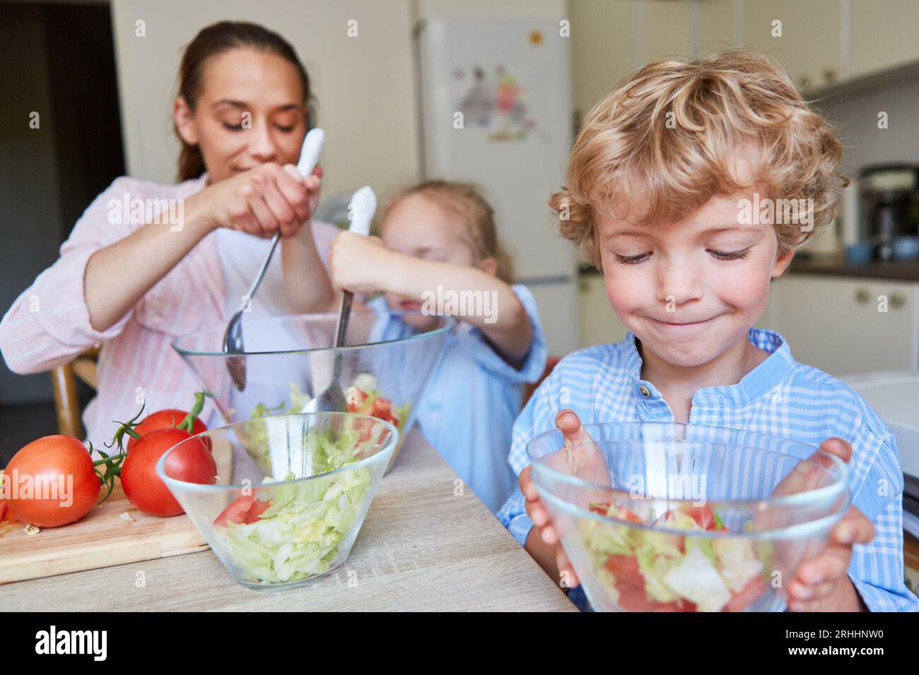 Smiling blond boy looking at salad bowl with mother and daughter making food at home Stock Photo