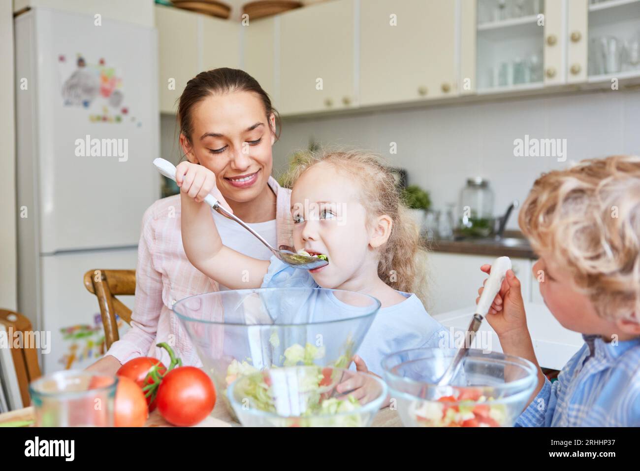 Daughter eating vegetable salad using spork with mother and brother in kitchen at home Stock Photo