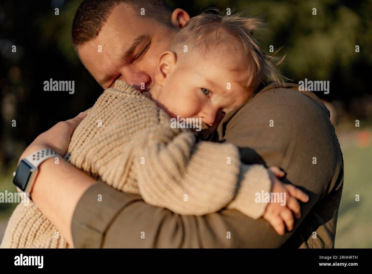 young dad with eyes closed hugging baby son putting head to his shoulder and sucking a pacifier. they are standing in park. High quality photo Stock Photo