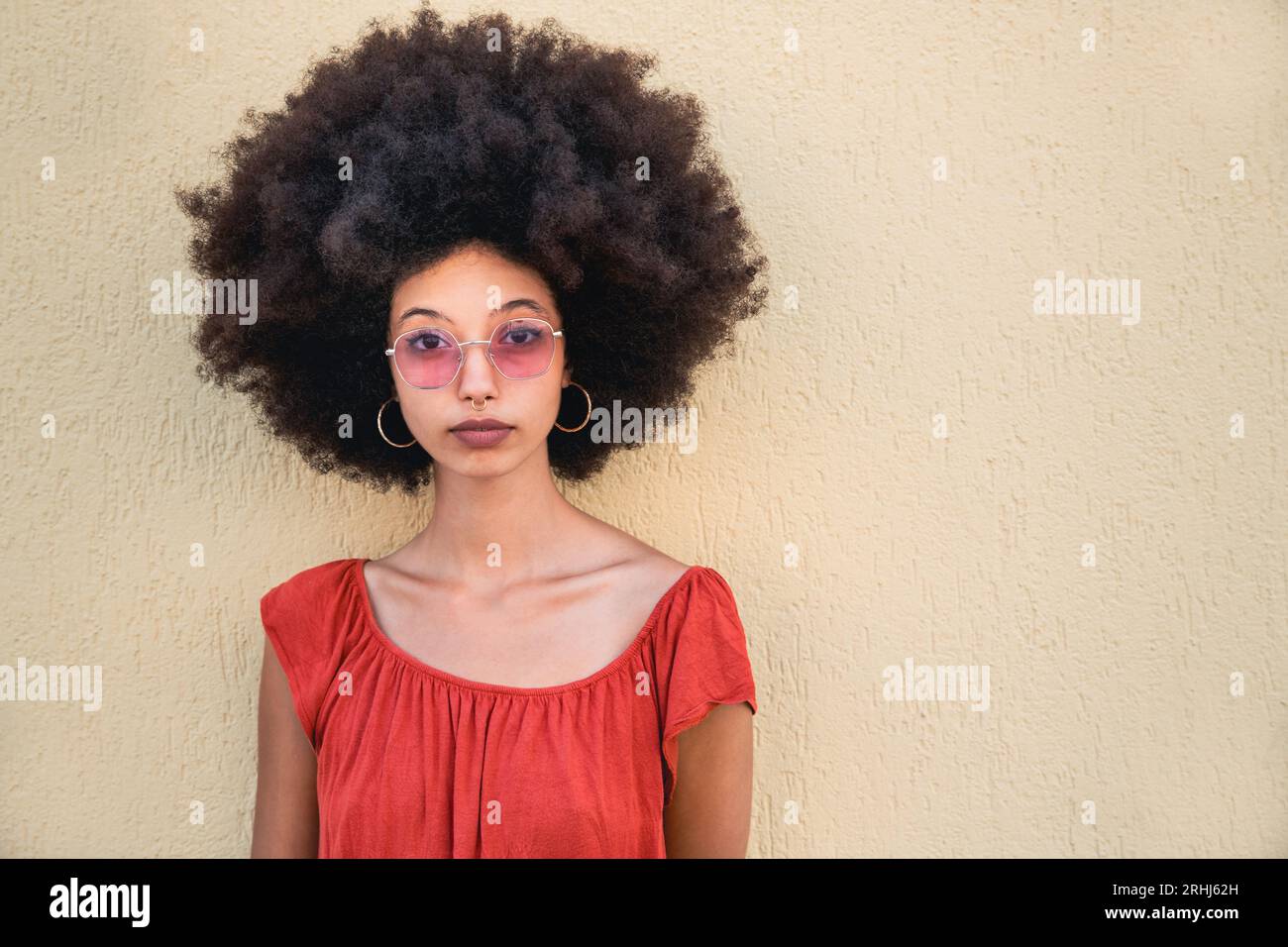 Portrait of serious young Moroccan female in sunglasses with Afro hair looking at camera while standing on street against beige wall in daylight Stock Photo