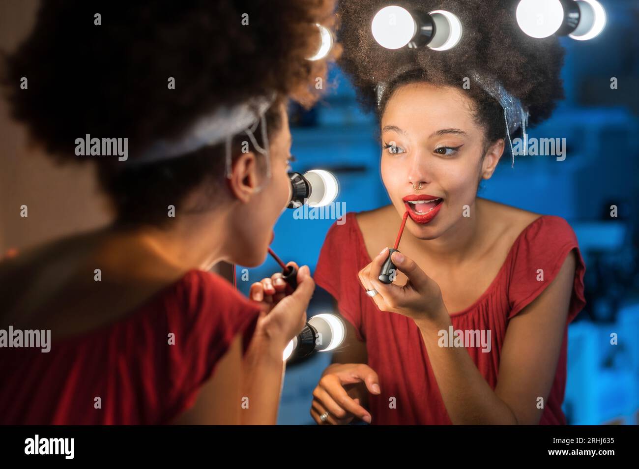 Young Moroccan female with Afro hair headband looking away while standing in front of makeup mirror with illuminated lights and putting red lipstick a Stock Photo