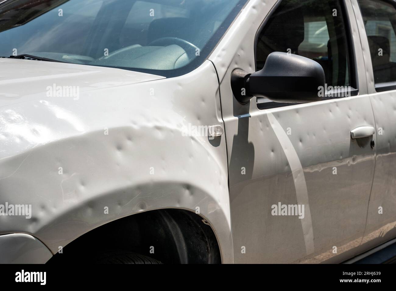 Damaged white car with many dents parked on city street on sunny day after hailstorm Stock Photo