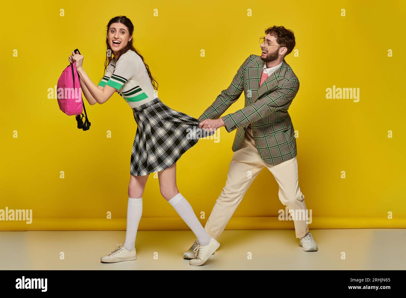 funny students, college life, happy man pulling skirt of female classmate on yellow backdrop, run Stock Photo