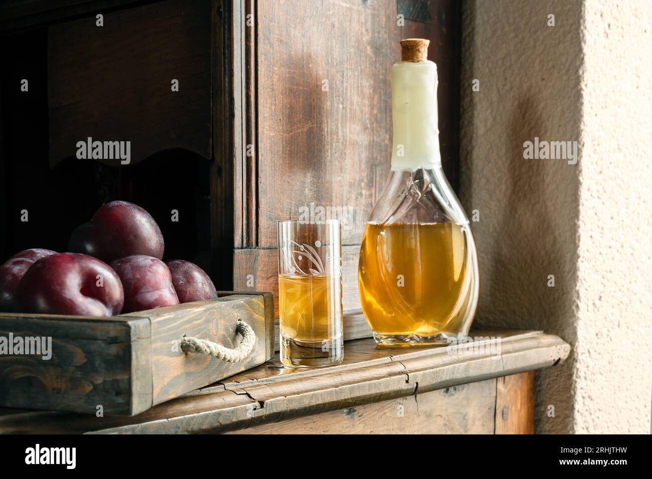 Traditional Balkan plum brandy - rakia slivovica or rakija in the bottle, a wineglass with sljivovica and fresh plums on the old sideboard in daylight Stock Photo