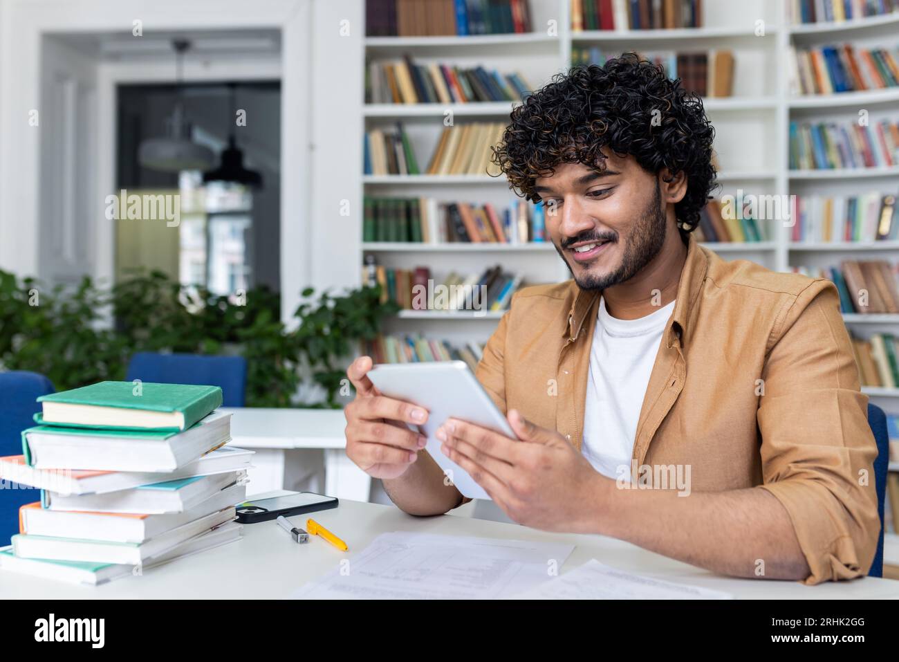 Young successful student studying sitting inside university academic library, man smiling holding tablet computer, Hispanic man using study app. Stock Photo