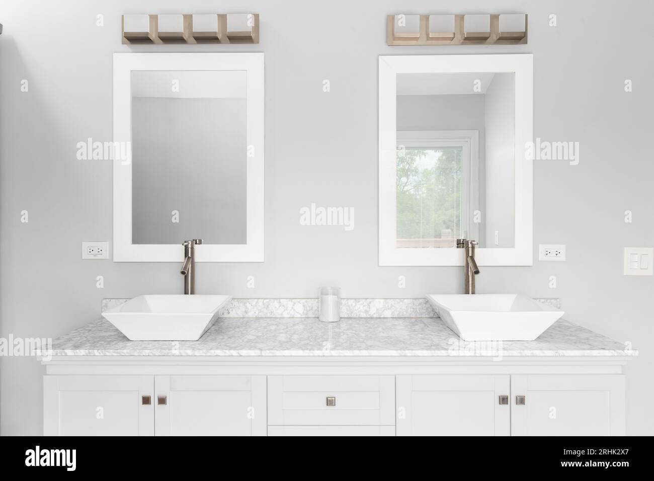 A bathroom detail with a white cabinet, two vessel sinks, marble countertop, and a bronze light fixture mounted above white framed mirrors. Stock Photo