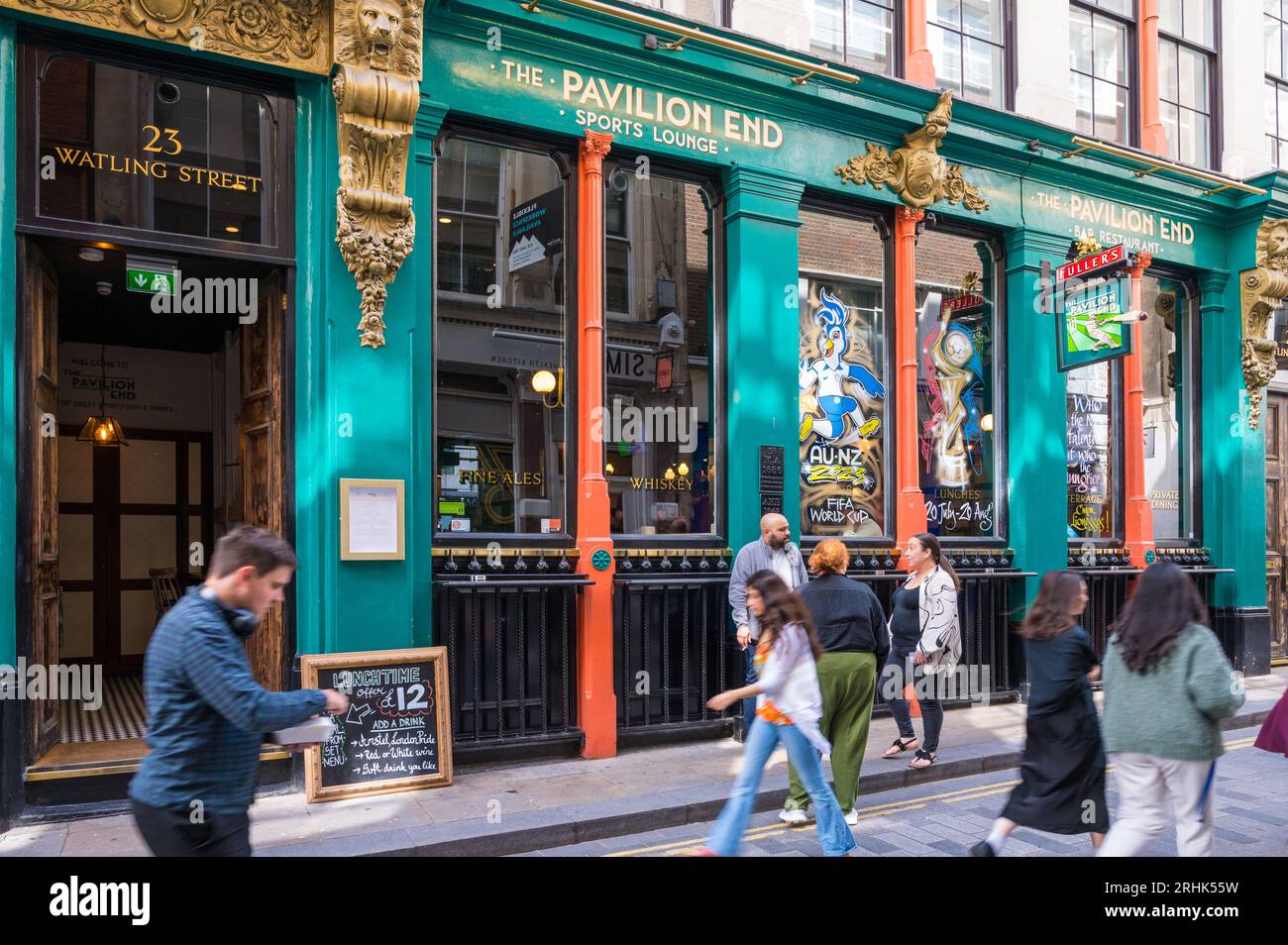 People stand in conversation and others pass by the colourful exterior of The Pavilion End, a Fuller's pub on Watling Street, City of London, England Stock Photo