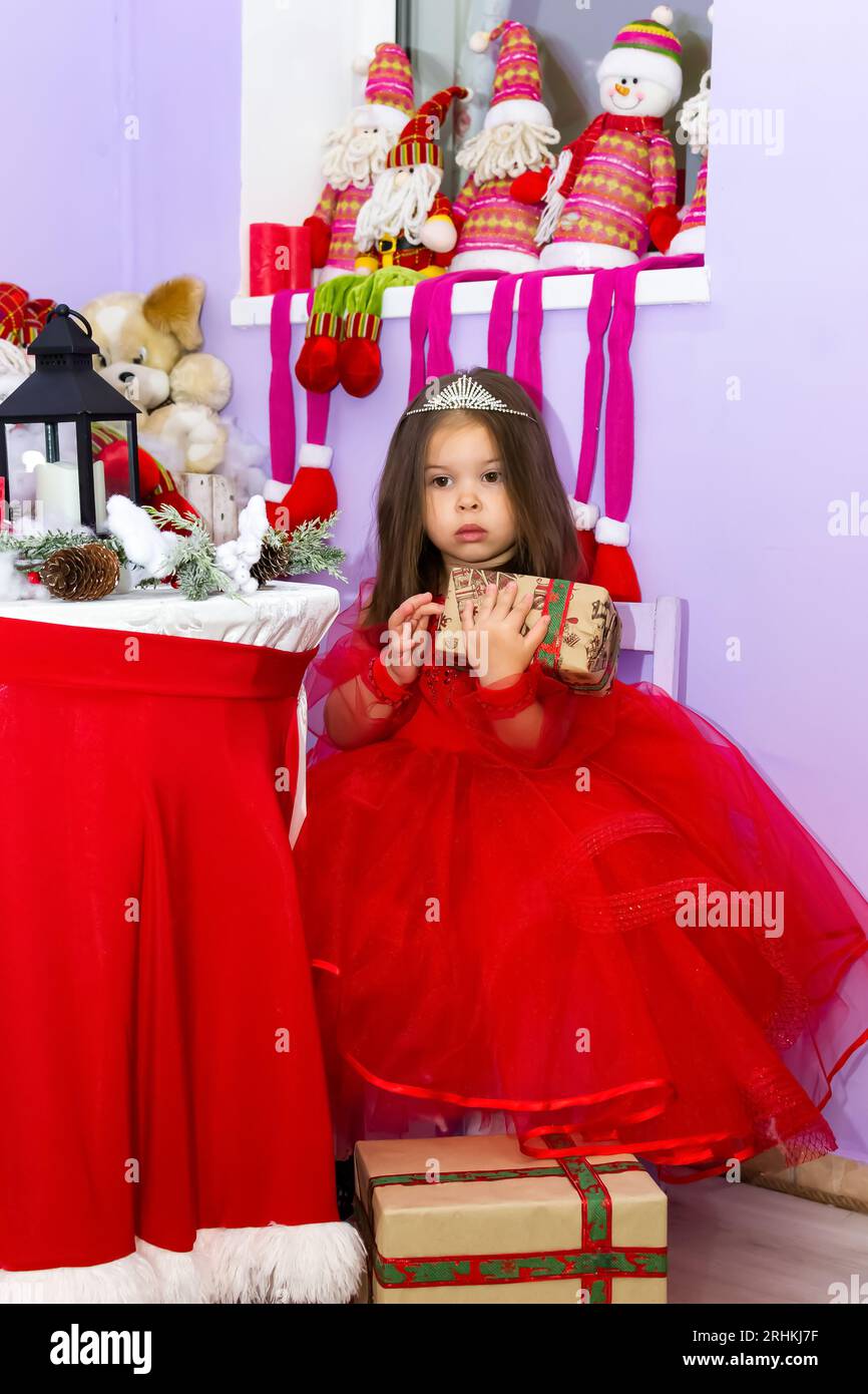 A little girl in a red dress is holding a gift. A little girl holds a gift on a chair. A child in a red dress holds a gift box. Girl looking at camera Stock Photo