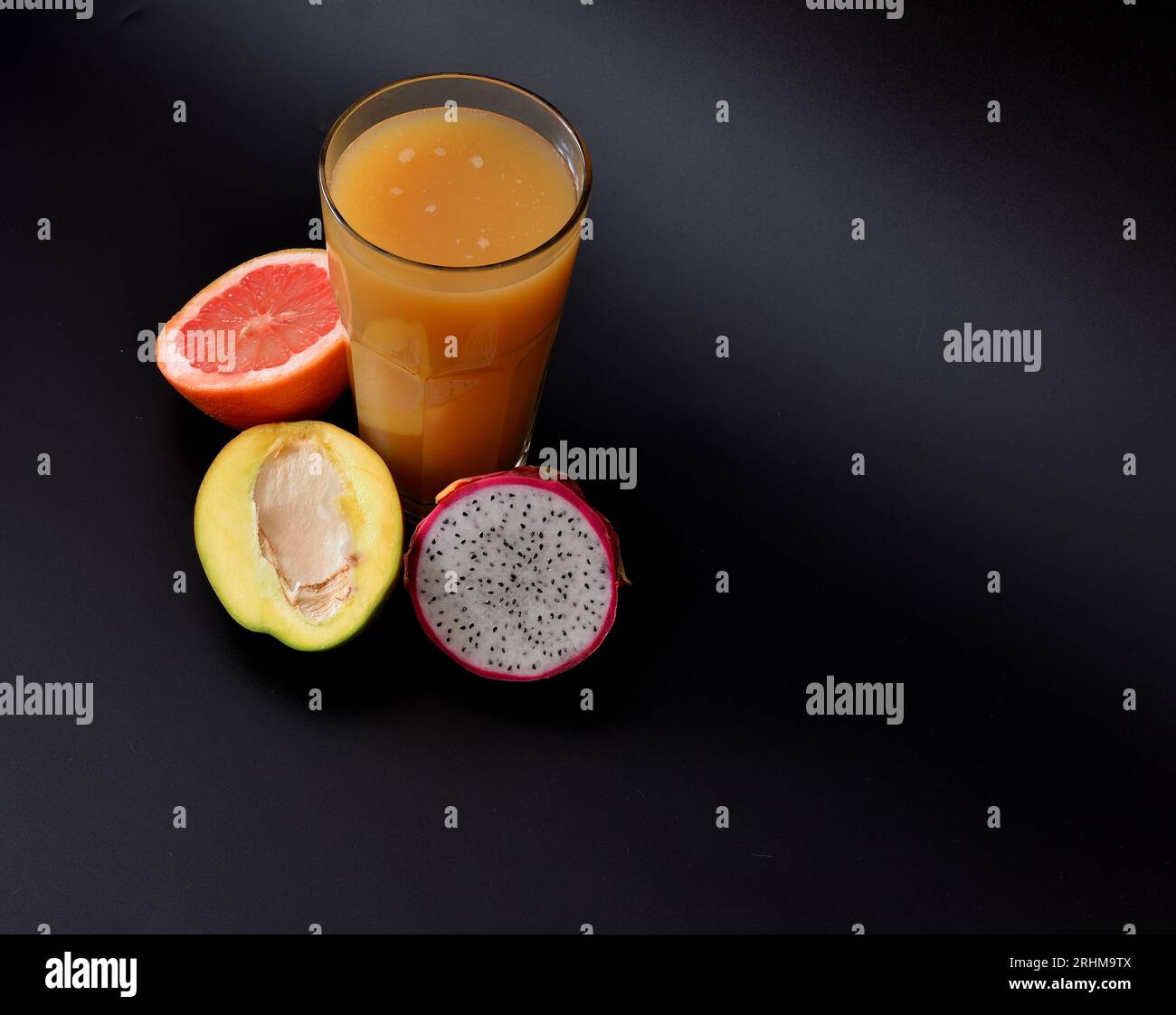A tall glass of freshly squeezed fruit juice on a black background, next to half a ripe mango, grapefruit and pitaya. Close-up. Stock Photo