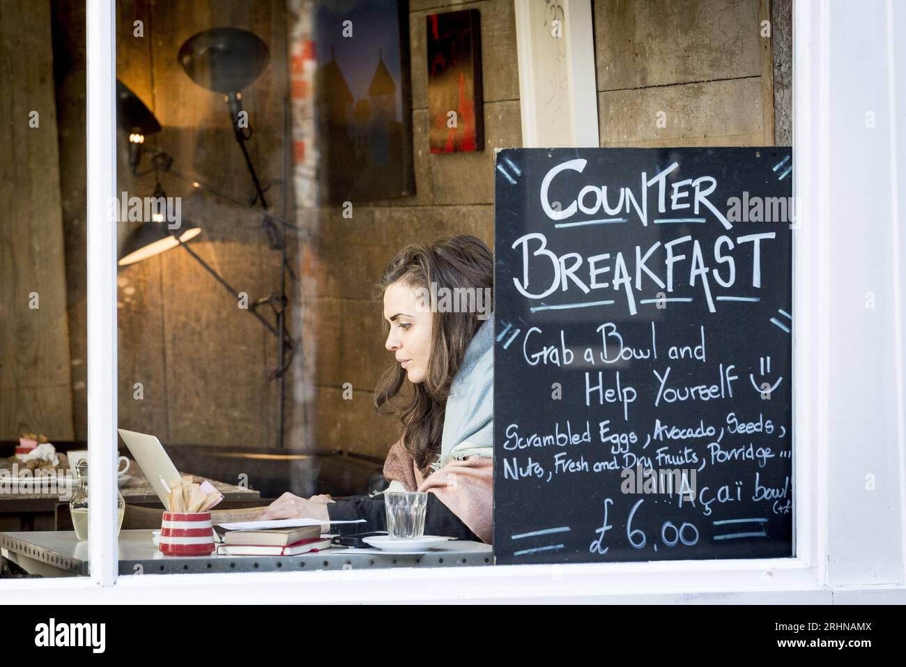 Woman works on laptop inside trendy Redchurch Street café in East London, with chalkboard menu offering healthy breakfast options Stock Photo