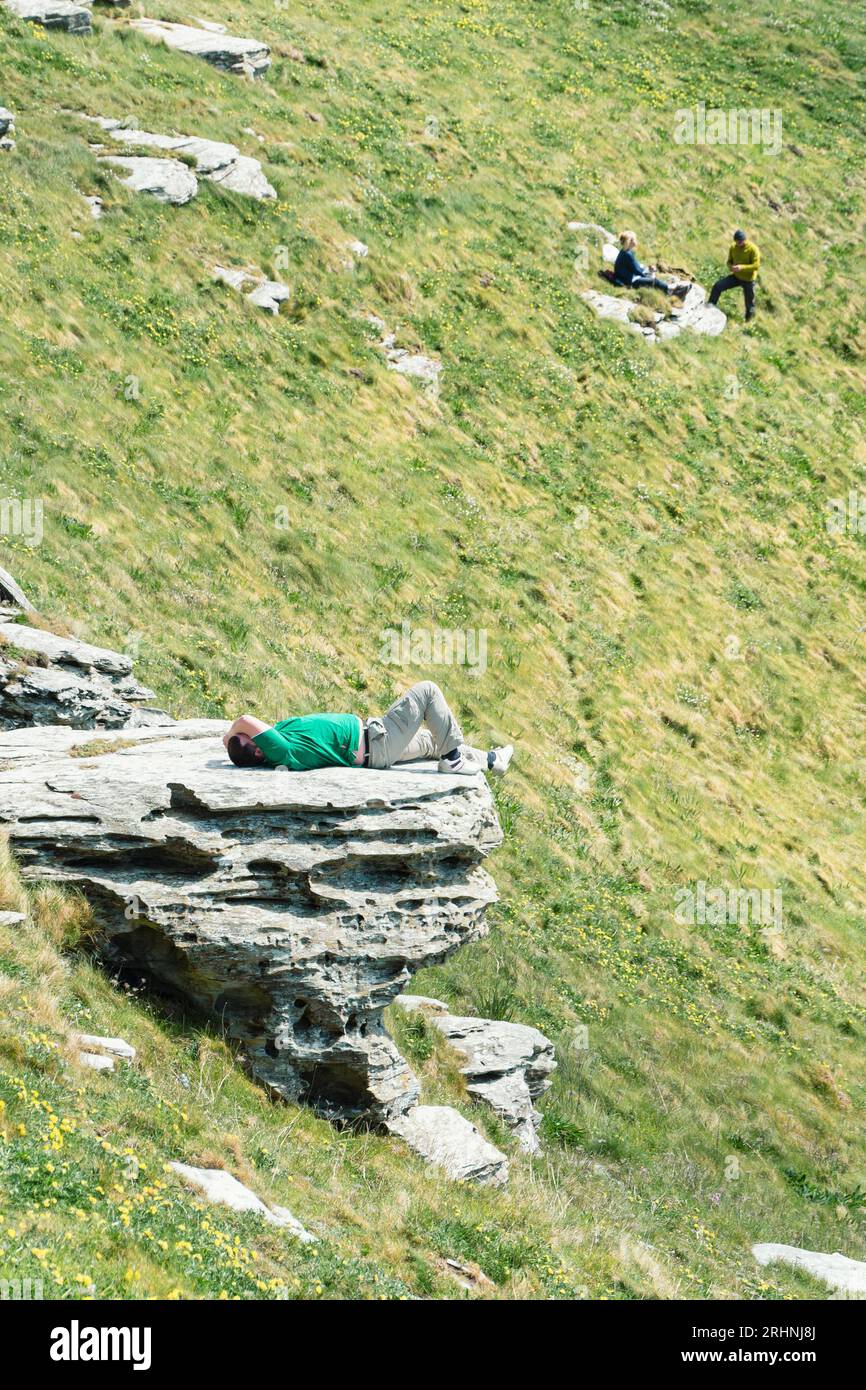 A man relaxes in the sun on a large flat rock, while a couple talk in the background in a sloping bank near Tintagel in Cornwall UK Stock Photo