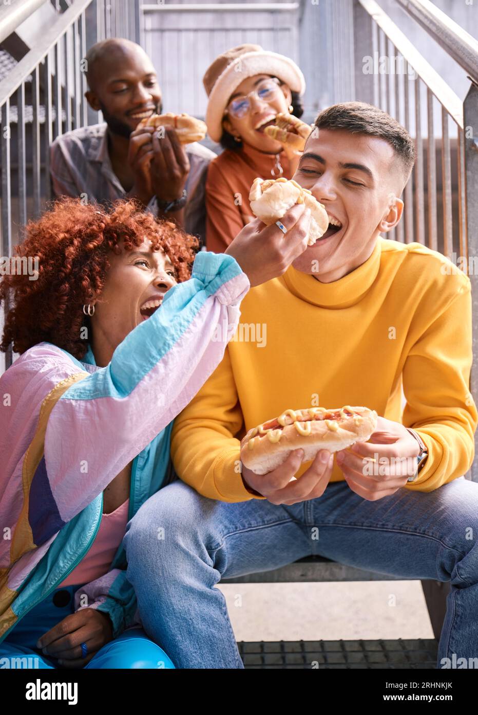 Friends, city and feeding food outdoor for happiness, funny laugh and fun on stairs. Diversity, eating and gen z group of men and women with a hotdog Stock Photo