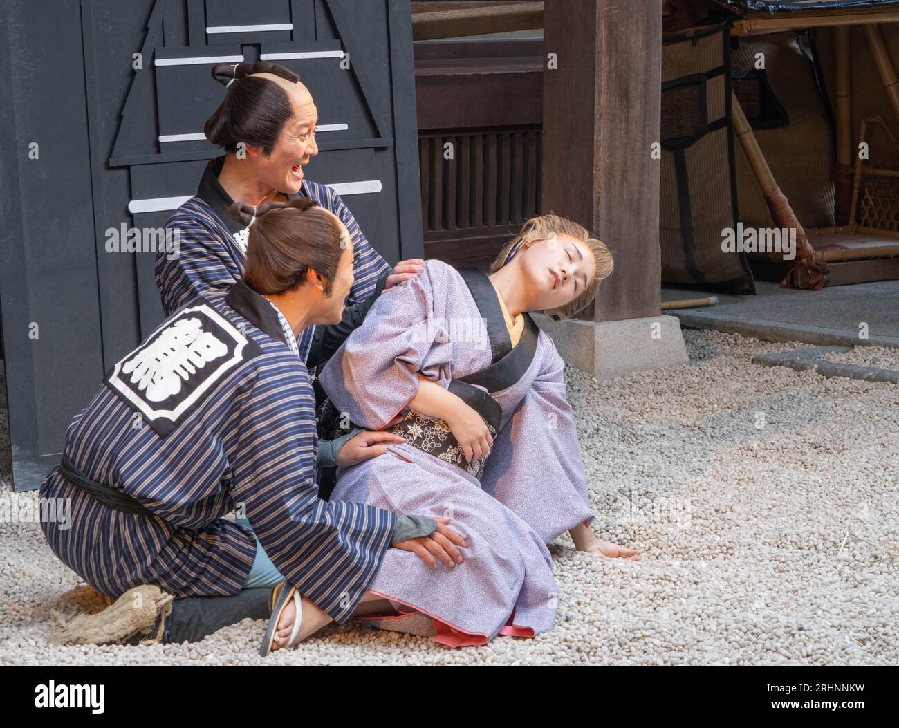 Two men dressed in period costume holding a dead woman wearing a yukata in an edo period comedy play in Edomura Wonderland. Stock Photo