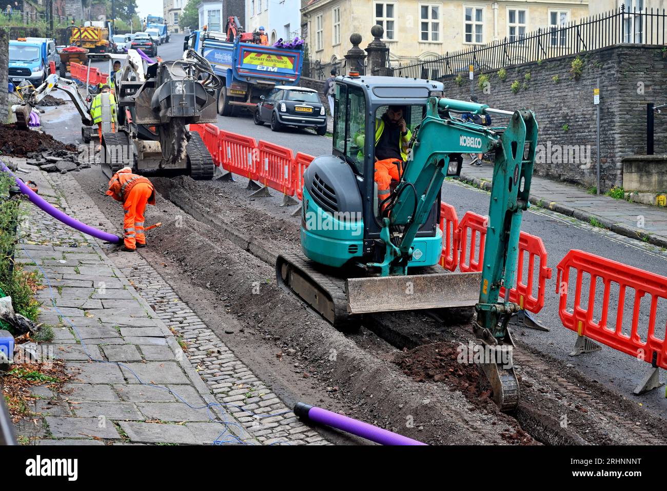 Trenching machine (Bobcat) used in conjunction with backhoe excavator digging trench in street for fibre optic duct in communications upgrade, Bristol Stock Photo