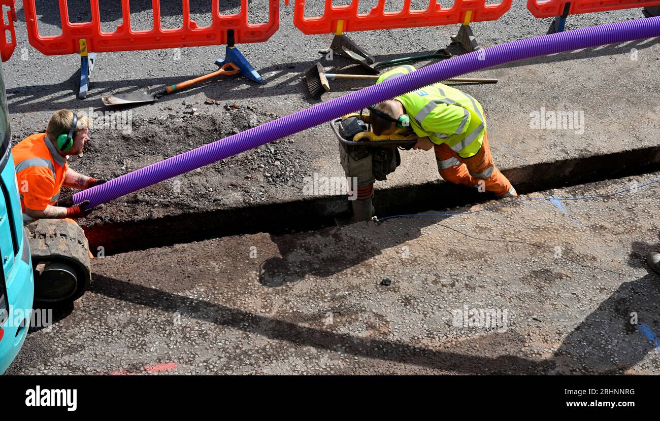 Workmen installing ducting for fibre optic cable in trench cut in road