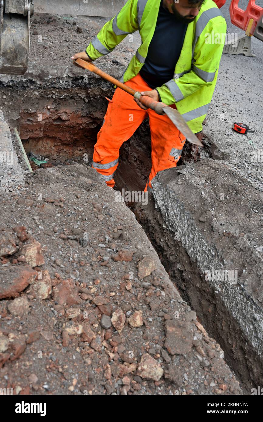 Workman cleaning hole for junction box in ducting for fibre optic cable in trench cut in road, UK Stock Photo