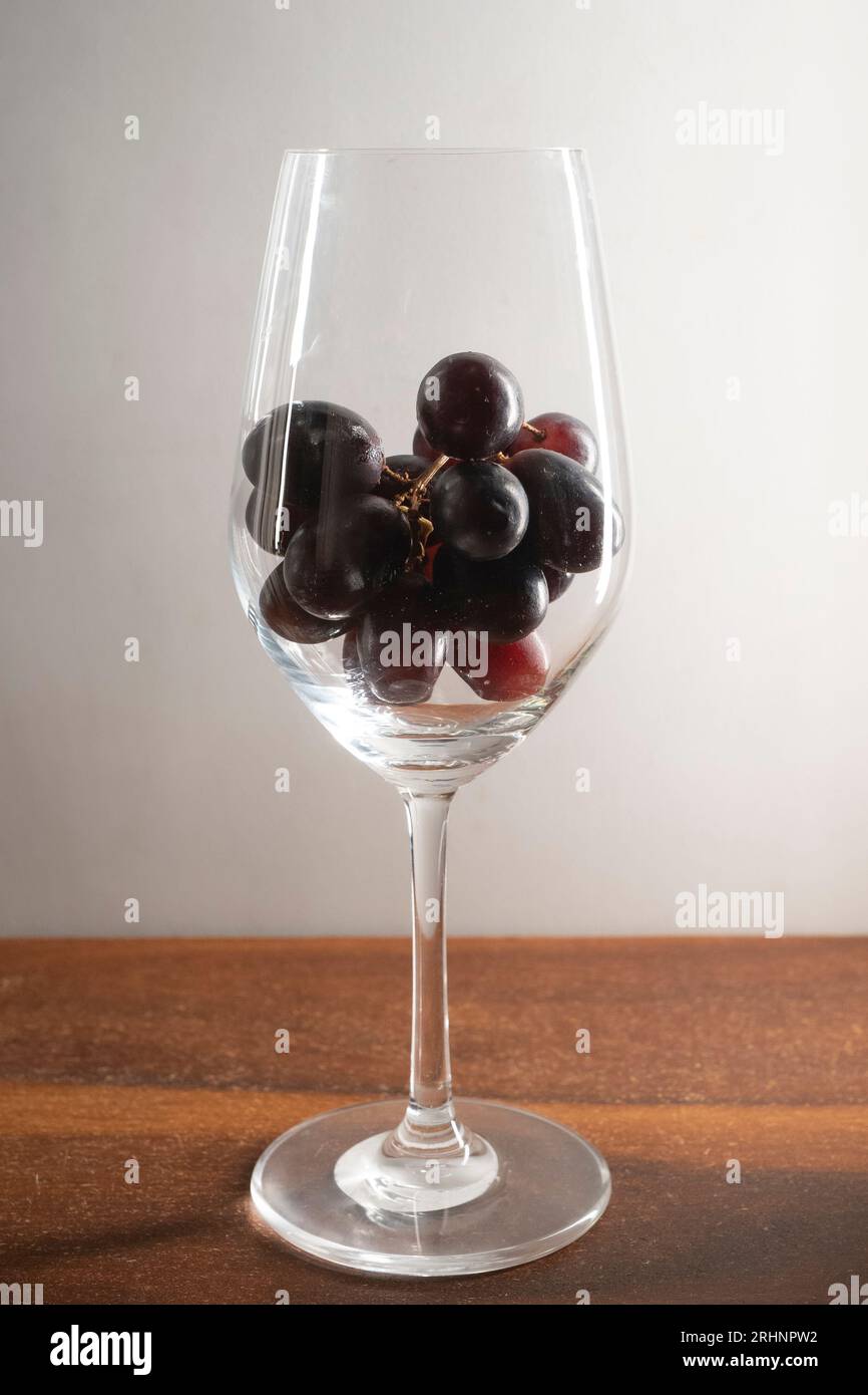 A bunch of dark red grapes in a wine glass kept on wooden table against white background Stock Photo