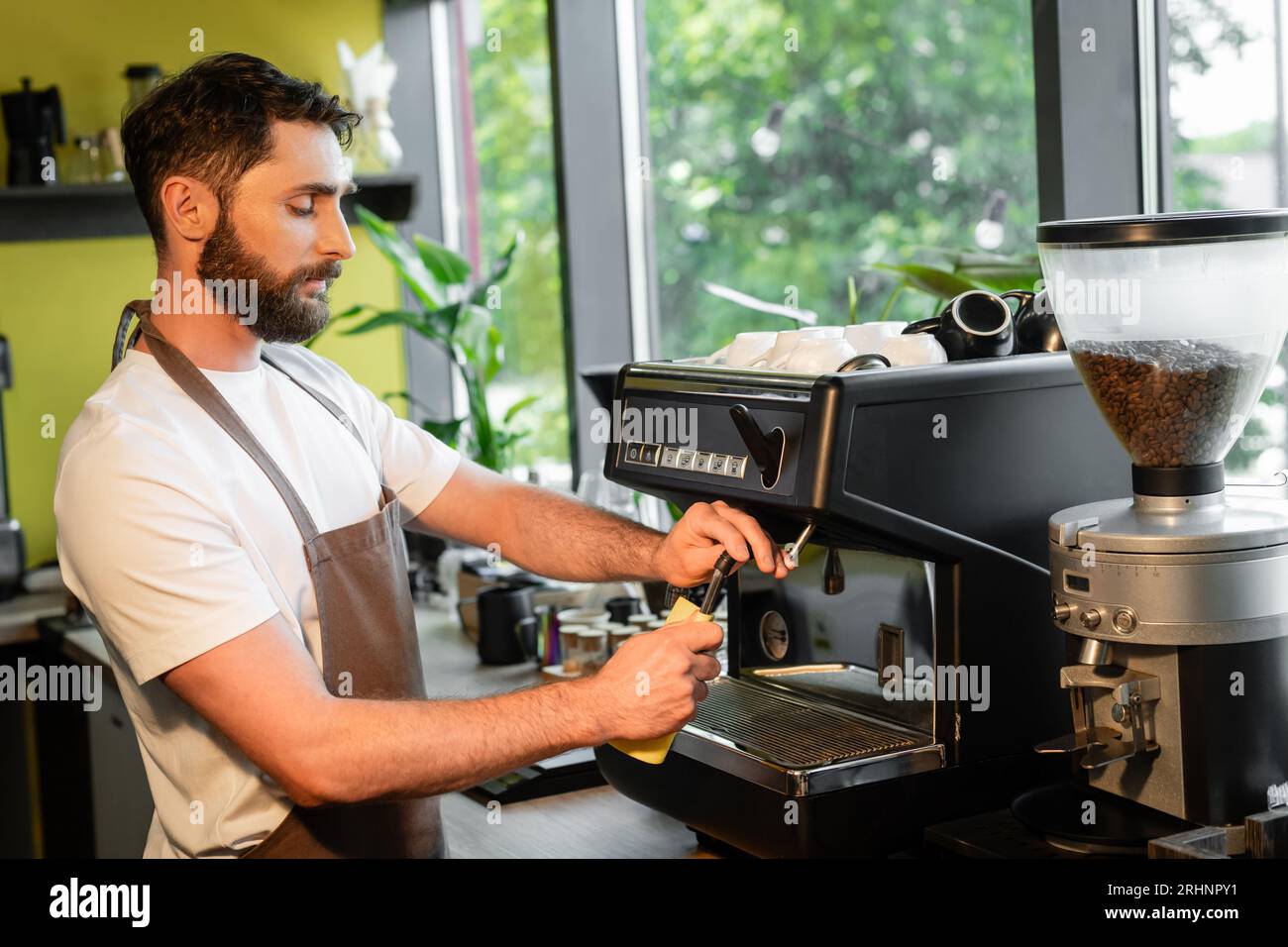 bearded barista cleaning coffee machine nozzle with rag while working in coffee shop Stock Photo