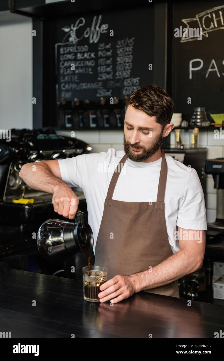 bearded barista pouring coffee from pot in glass with ice cubes, bar in coffee shop, ice coffee