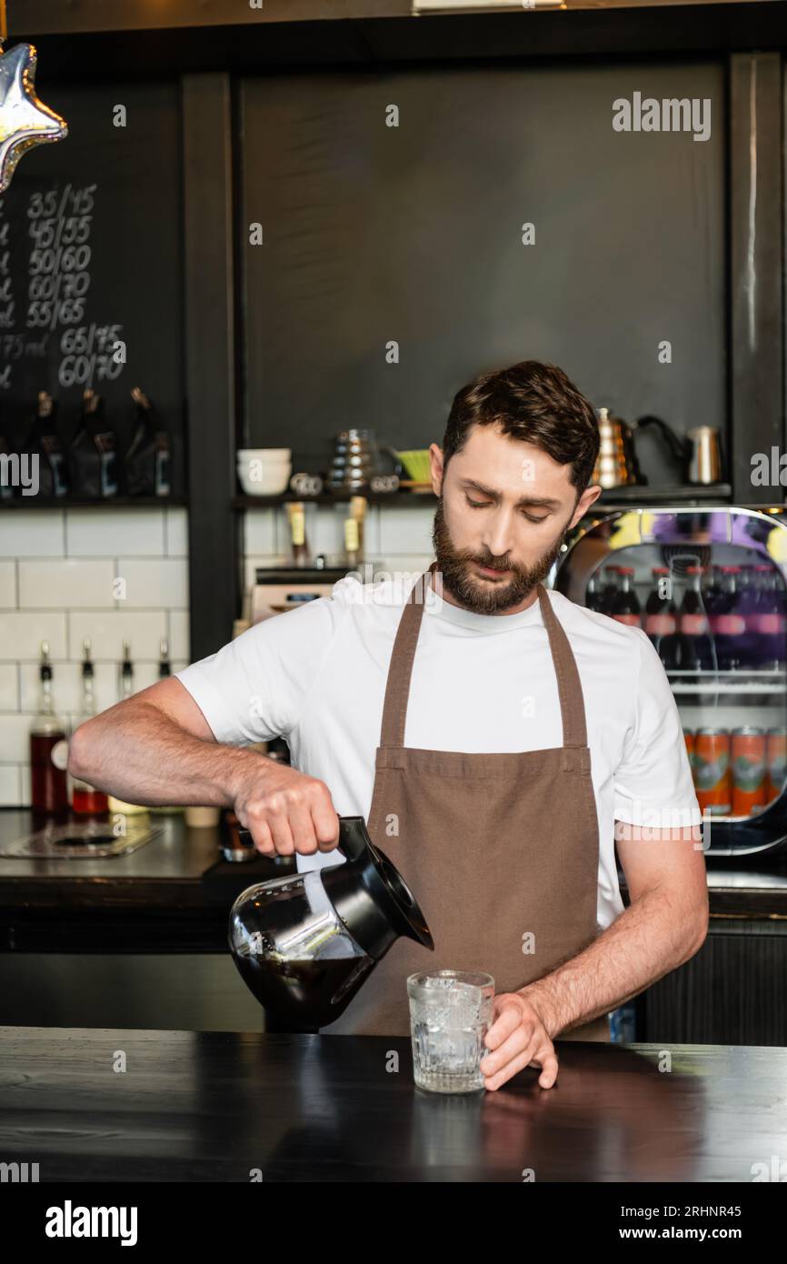 barista in apron pouring coffee from pot in glass with ice cubes on bar in coffee shop Stock